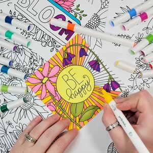 May include: A close-up of a colouring page with the words "Be Happy" in a yellow circle surrounded by flowers and sun rays. A hand is holding a yellow marker over the page.