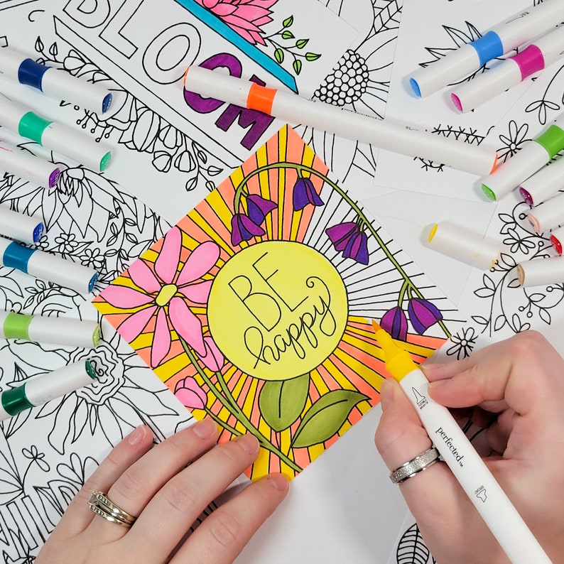 May include: A close-up of a colouring page with the words "Be Happy" in a yellow circle surrounded by flowers and sun rays. A hand is holding a yellow marker over the page.
