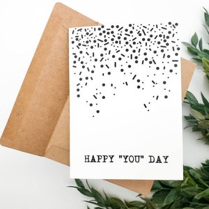 May include: A white card with black confetti and the text "HAPPY "YOU" DAY" printed on it. The card is sitting on a brown envelope.