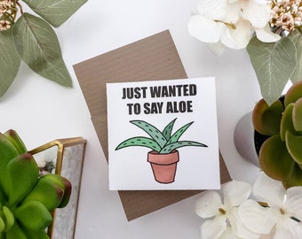 May include: A white card with a brown envelope behind it. The card has a cartoon drawing of an aloe plant in a pot and the text "Just wanted to say aloe".