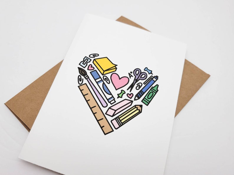 May include: A white card with a heart shape made of colorful school supplies, including a pencil, ruler, scissors, crayons, and a paintbrush. The heart is surrounded by a brown envelope.