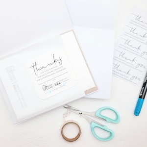 May include: A white card with a thank you message for a purchase, encouraging creativity and supporting small businesses. The card is in a clear plastic sleeve. There is a roll of tape, a pair of scissors, and a blue marker on a white surface.