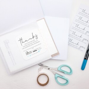 May include: A white card with the text "thanks" and a message thanking the customer for their purchase, encouraging creativity, and supporting small businesses. The card is on a white surface with a blue marker, scissors, and a roll of washi tape.