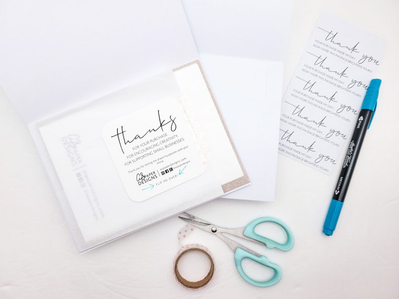 May include: A white card with the text "thanks" and a message thanking the customer for their purchase, encouraging creativity, and supporting small businesses. The card is on a white surface with a blue marker, scissors, and a roll of washi tape.
