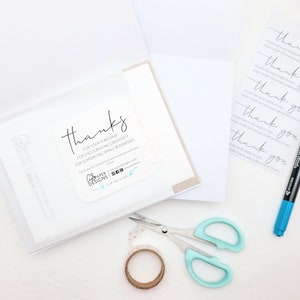 May include: A white card with a thank you message for a purchase, encouraging creativity and supporting small businesses. The card is in a clear plastic sleeve. There is a roll of tape, a pair of scissors, and a blue marker on a white surface.