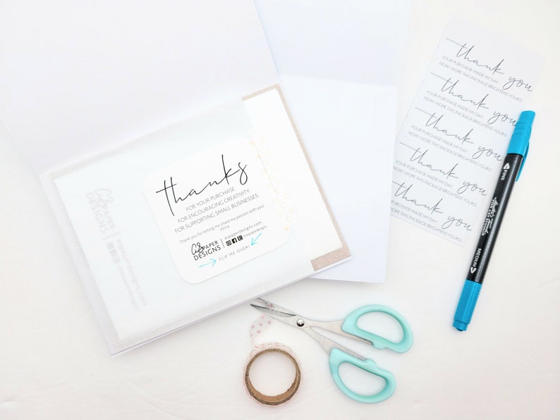 May include: A white card with a thank you message for a purchase, encouraging creativity and supporting small businesses. The card is in a clear plastic sleeve. There is a roll of tape, a pair of scissors, and a blue marker on a white surface.