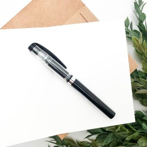 May include: A blank white card with a black pen lying on top of it. The card is on a brown paper envelope and a green leafy branch is in the background.