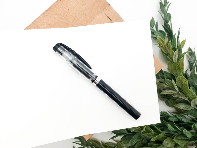 May include: A blank white card with a black pen lying on top of it. The card is on a brown paper envelope and a green leafy branch is in the background.