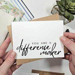 May include: A white greeting card with the message "YOU ARE A difference maker" in black script. The card is inside a brown envelope. Other stationery and a succulent plant are in the background.