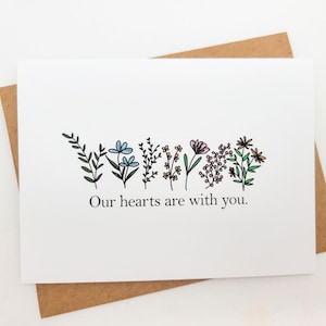 May include: A white card with a line of hand-drawn flowers in shades of blue, pink, and green. The text "Our hearts are with you." is printed below the flowers.