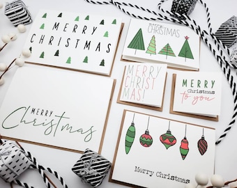 May include: A collection of Christmas greeting cards with various designs. Some cards feature the text "Merry Christmas" in different fonts, while others display green Christmas trees or colorful ornaments. The cards are on a white surface with decorative elements.