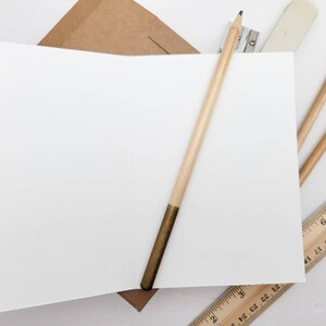 May include: A white sheet of paper with a brown pencil, a pencil sharpener, a ruler, and two wooden pencils on a white surface.