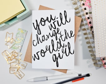 May include: A white card with black lettering that reads "you will change the world, girl". The card is sitting on a brown envelope and a white surface.