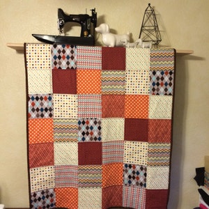 May include: A patchwork quilt with a variety of patterns and colors, including orange, brown, white, and gray. The quilt is made up of squares and is hanging on a wall.