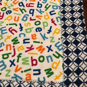 May include: A white fabric with a blue border. The fabric is covered in colorful letters of the alphabet. The border is a repeating pattern of blue circles on a white background.
