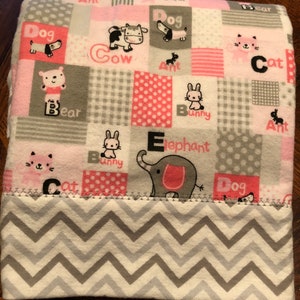 May include: A pink and gray baby blanket with a patchwork design featuring various animals and letters. The blanket has a chevron pattern on the back.
