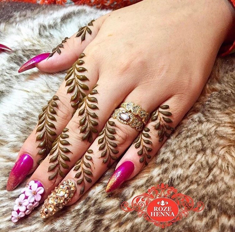 May include: A hand with henna designs on the fingers, featuring a leafy pattern. The nails are painted a bright pink and adorned with rhinestones and white flowers.