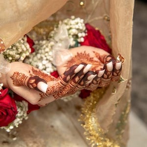 May include: Close-up of a bride's hands with intricate henna designs, adorned with white nail polish. The hands are holding a bouquet of white flowers and red roses, partially covered by a sheer beige veil.