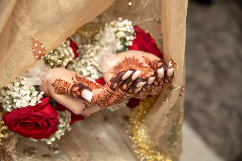 May include: Close-up of a bride's hands with intricate henna designs, adorned with white nail polish. The hands are holding a bouquet of white flowers and red roses, partially covered by a sheer beige veil.