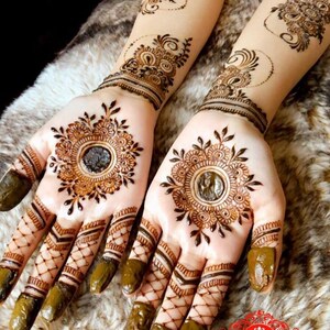 May include: Intricate henna designs on both hands, featuring a circular pattern with a smaller circle in the center. The fingers are decorated with a lattice pattern and the design extends up the forearms.