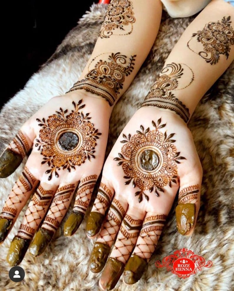 May include: Intricate henna designs on both hands, featuring a circular pattern with a smaller circle in the center. The fingers are decorated with a lattice pattern and the design extends up the forearms.