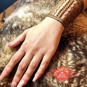 May include: A close-up of a henna tattoo on a person's wrist. The tattoo is a detailed design with a floral pattern and a row of small dots. The tattoo is a dark brown color and is on a light brown background.