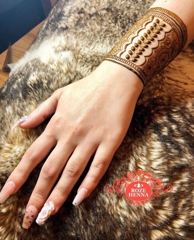 May include: A close-up of a henna tattoo on a person's wrist. The tattoo is a detailed design with a floral pattern and a row of small dots. The tattoo is a dark brown color and is on a light brown background.