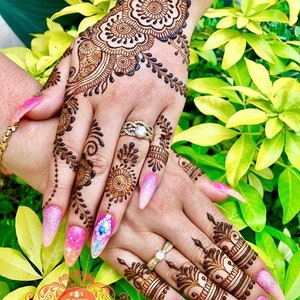 May include: A woman's hands with intricate henna designs on the back of her hand and fingers. Her nails are painted with a gradient of pink and white with glitter and rhinestones.