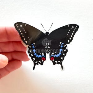 Puede incluir: Una pegatina de mariposa negra con lunares blancos en las puntas de las alas y detalles azules y rojos. La pegatina está sujeta por una mano sobre un fondo blanco. El texto "BREANN COOK" es visible.