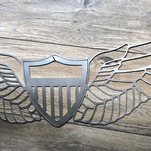 May include: A metal wall decor featuring a stylized American shield with wings. The shield has a classic design with vertical stripes and a star at the top. The wings are spread wide and have a detailed, layered design.