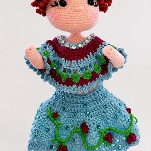 Puede incluir: Muñeca de ganchillo hecha a mano con cabello rizado rojo y tez rosa claro. La muñeca viste un vestido azul con enredaderas verdes y rosas rojas, y una parte superior azul y burdeos con detalles de joyas. Una flor blanca está en el cabello de la muñeca.