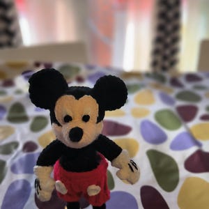 Puede incluir: Una figura de Mickey Mouse hecha a mano con ganchillo. La figura tiene una cabeza negra con orejas grandes, una cara beige, brazos negros, guantes blancos, pantalones cortos rojos y zapatos amarillos. La figura está sobre una superficie estampada.