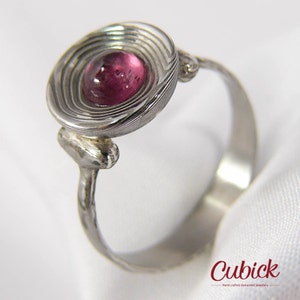 May include: A silver ring with a circular, layered design around a round, pink gemstone. The ring band is smooth and simple, with a textured connection to the gemstone setting. The ring is set against a white background.