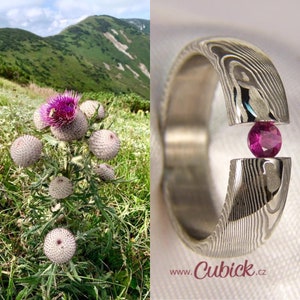 May include: A split silver ring with a patterned design, holding a round, deep pink gemstone. The ring is set against a white background. The image also shows a field of purple thistle flowers and a mountain range.