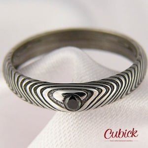 Pode incluir: Um anel de prata com um design de padrão de ondas único e uma pequena pedra preta. O anel tem uma aparência escura e texturizada, sobre um fundo branco. O logotipo Cubick é visível.