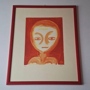 Op de afbeelding: Abstracte kunstprint met een gestileerde menselijke figuur in oranje en rode tinten. Het kunstwerk is ingelijst in een rode houten lijst, met een crèmekleurige passe-partout. De print heeft een minimalistische stijl en is rechtsonder gesigneerd.