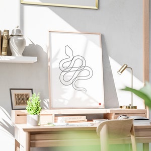May include: A minimalist black line drawing of a snake on a white framed print. The artwork is displayed on a wooden desk with a small plant, books, and a gold desk lamp. The room has a neutral colour palette and a wooden desk.