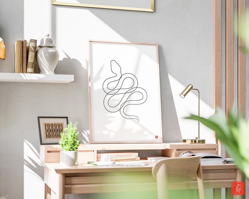 May include: A minimalist black line drawing of a snake on a white framed print. The artwork is displayed on a wooden desk with a small plant, books, and a gold desk lamp. The room has a neutral colour palette and a wooden desk.