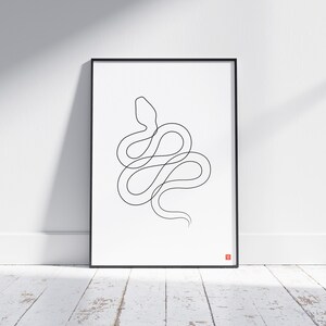 May include: A framed art print with a minimalist black line drawing of a snake on a white background. The artwork is in a black frame and leans against a white wall. A small red square is in the bottom right corner.