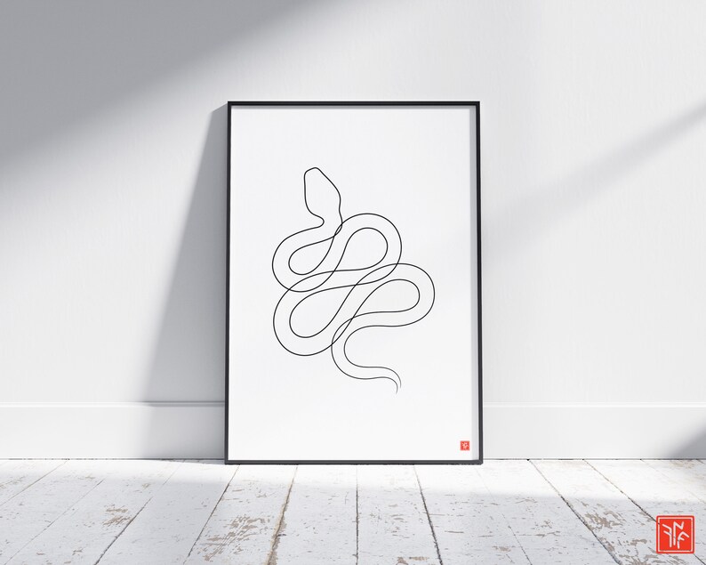May include: A framed art print with a minimalist black line drawing of a snake on a white background. The artwork is in a black frame and leans against a white wall. A small red square is in the bottom right corner.