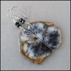 May include: Dangle earrings featuring natural stone slices with a unique pattern of black, white, and brown hues. Each earring has a silver hook and is accented with three small black beads. The earrings are displayed on a neutral gray surface.