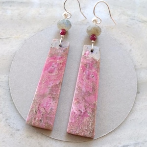 May include: A pair of dangle earrings featuring long, rectangular pink and brown stone pendants. Each earring is suspended from a silver-toned hook, accented with a small, faceted grey bead and a tiny, round, deep red bead above the stone.
