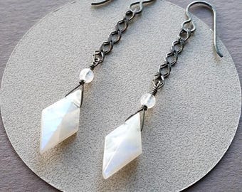 May include: A pair of dangle earrings featuring a diamond-shaped, faceted white gemstone. The earrings have a dark silver chain and hook, with a small, round white bead above the gemstone. The earrings are displayed on a light gray surface.
