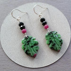 May include: A pair of earrings featuring leaf-shaped green stones with black inclusions and pink accents. The earrings have silver hooks and are adorned with black and pink beads. The earrings are displayed on a light gray surface.