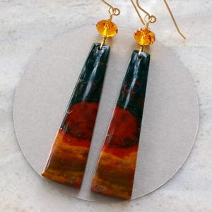 May include: A pair of dangle earrings featuring long, flat, trapezoidal-shaped stones with a polished finish. The stones display a gradient of dark green, deep red, and earthy brown tones. Each earring is topped with a small, faceted amber-colored crystal bead and a gold-toned wire hook.