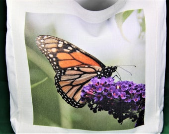 Puede incluir: Bolso tote blanco con una imagen de una mariposa monarca con alas naranjas, negras y blancas, posada sobre un racimo de pequeñas flores moradas. El fondo es verde, lo que sugiere un entorno natural.