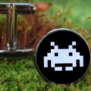 May include: A pair of silver cufflinks with a black background and a white pixelated alien graphic.