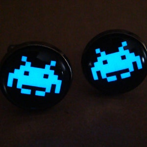 May include: A pair of cufflinks with a black background and a glowing blue space invader design.