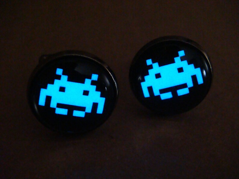 May include: A pair of cufflinks with a black background and a glowing blue space invader design.
