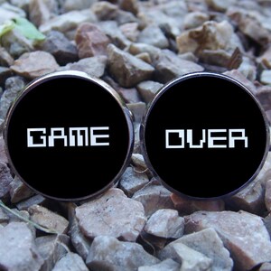 May include: A pair of black cufflinks with silver trim. The cufflinks feature the words "GAME OVER" in white pixelated text.